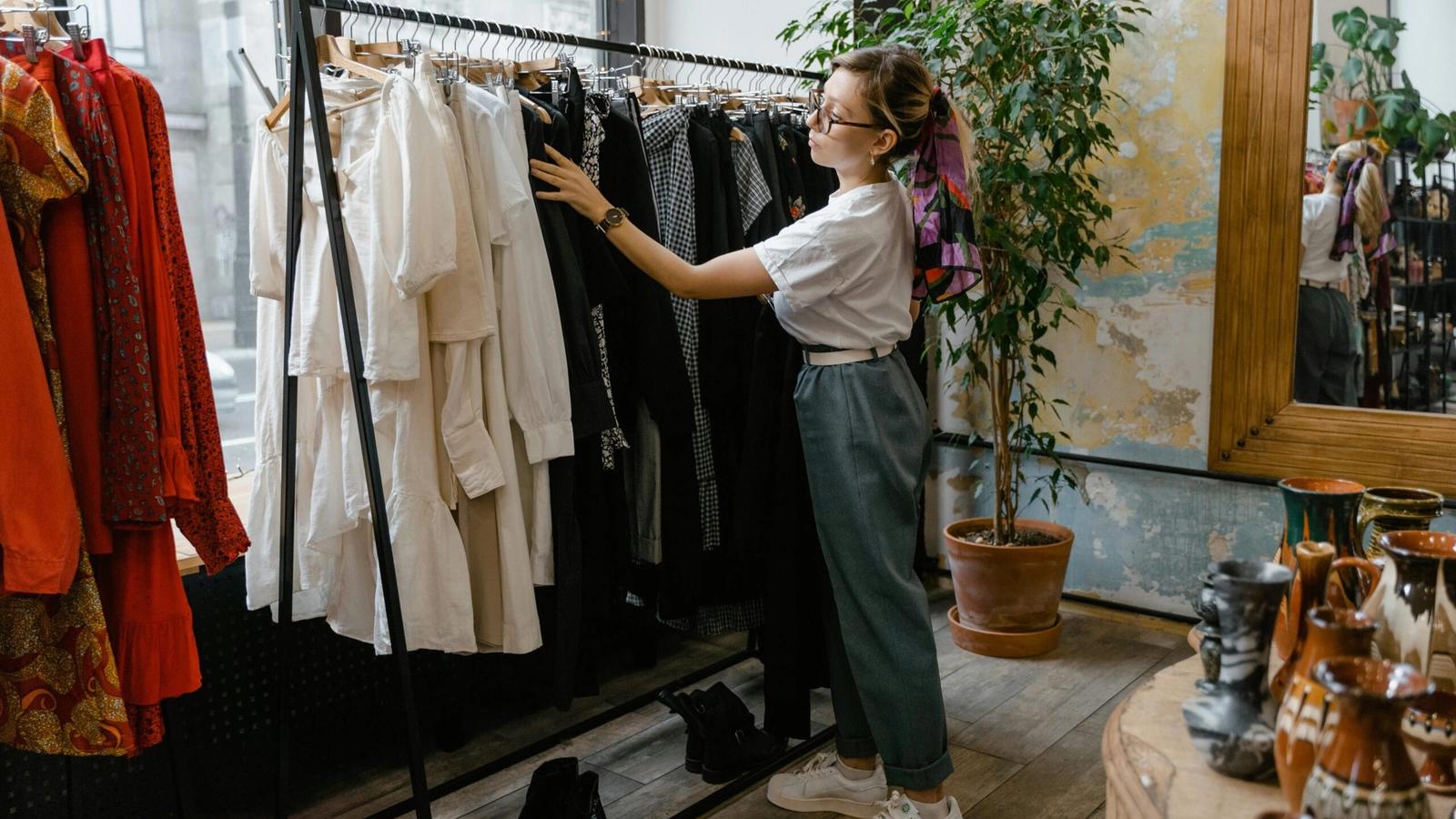 A woman wearing glasses, a white shirt, and jeans browses through clothes on a rack while shopping in Gatlinburg. The boutique features various garments, plants, and decorative items throughout the shop.