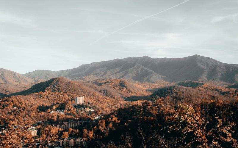 A scenic view of forested mountains with autumn foliage, scattered buildings, and a tall hotel structure nestled among the hills under a partly cloudy sky—one of Gatlinburg’s must see stops on any road trip.