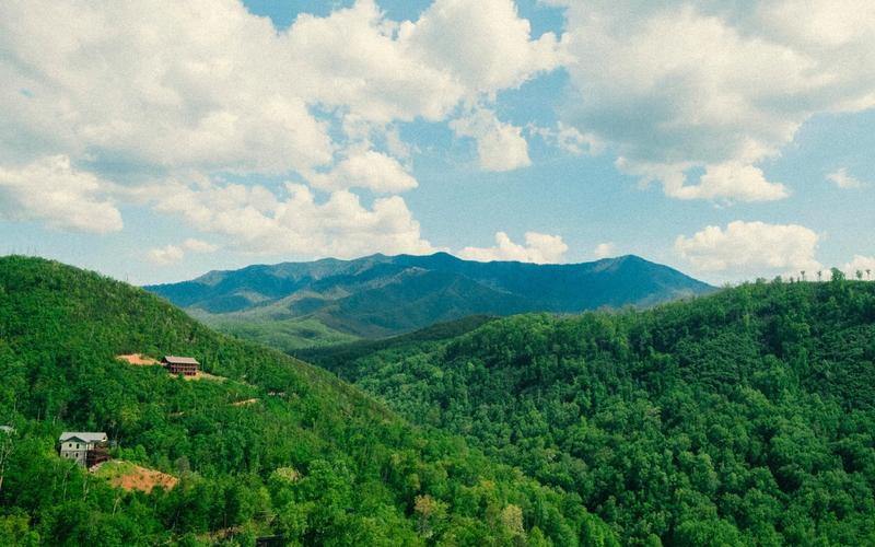 A scenic view of lush green forested hills near Gatlinburg, Tennessee, with a few houses scattered, set against a distant range of blue mountains under a partly cloudy sky.