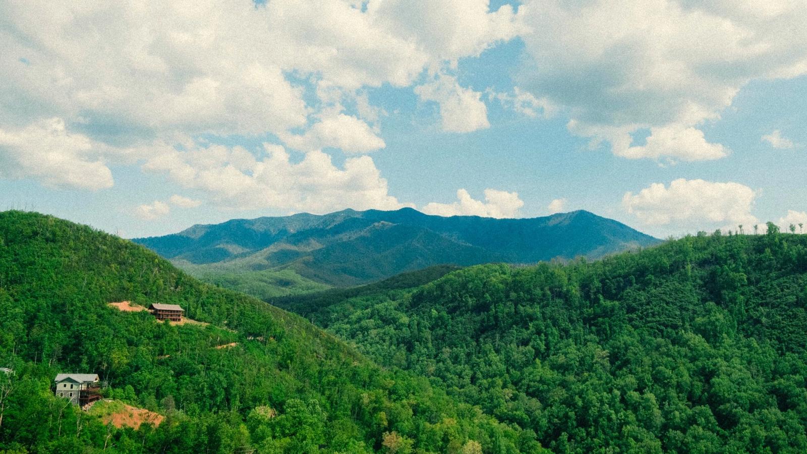 A scenic view of lush green forested hills near Gatlinburg, Tennessee, with a few houses scattered, set against a distant range of blue mountains under a partly cloudy sky.