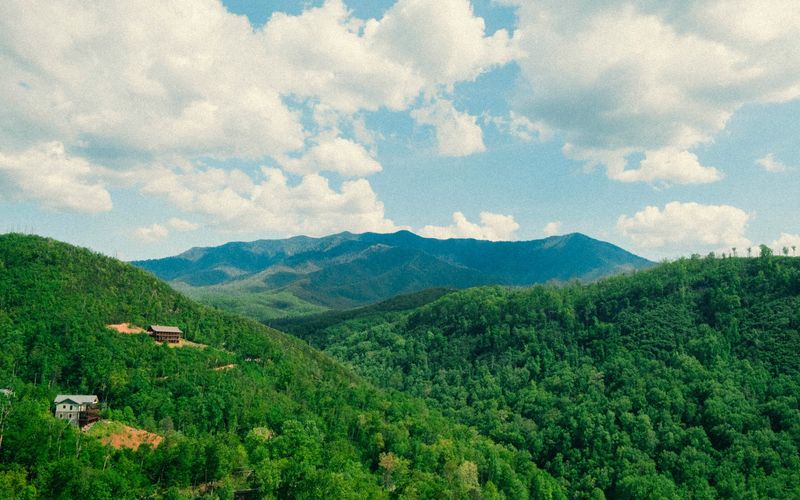 A scenic view of lush green forested hills near Gatlinburg, Tennessee, with a few houses scattered, set against a distant range of blue mountains under a partly cloudy sky.