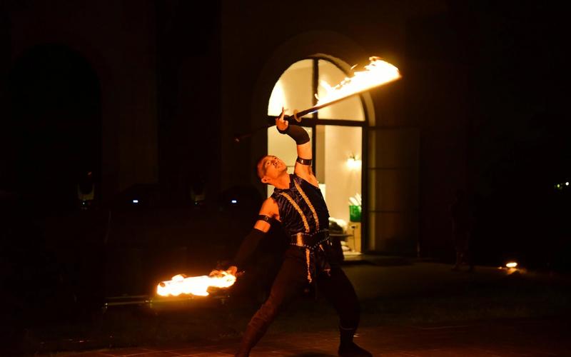 A fire performer in dark clothing dramatically poses at night, holding two flaming batons—one above his head and one extended outward—against the backdrop of an arched doorway, reminiscent of the thrilling Dinner Shows in Pigeon Forge.