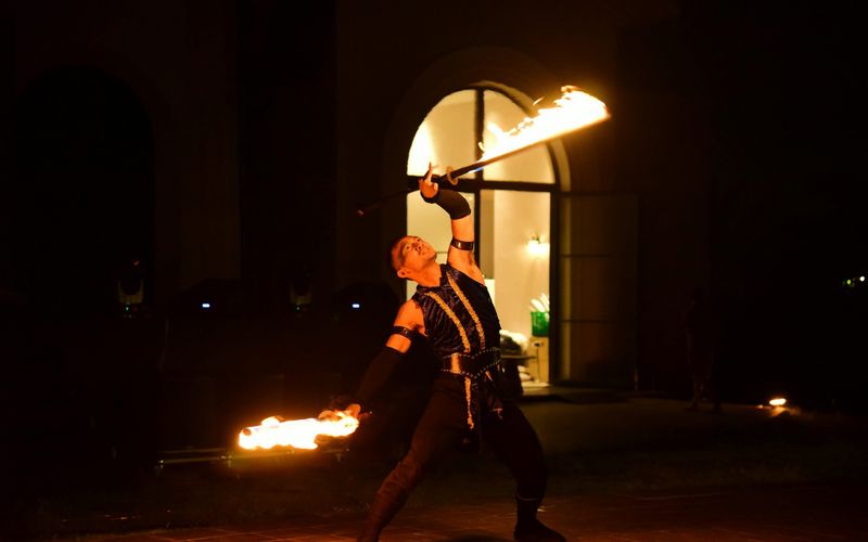 A fire performer in dark clothing dramatically poses at night, holding two flaming batons—one above his head and one extended outward—against the backdrop of an arched doorway, reminiscent of the thrilling Dinner Shows in Pigeon Forge.