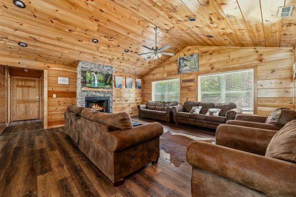 Spacious wooden living room with vaulted ceiling, brown sofas, animal hide rug, stone fireplace with fire, landscape photos on walls, and large windows letting in natural light—this inviting space evokes the warmth of a classic Smoky Mountain Cabin nestled within vibrant cabin communities.