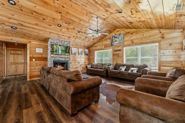 Spacious wooden living room with vaulted ceiling, brown sofas, animal hide rug, stone fireplace with fire, landscape photos on walls, and large windows letting in natural light—this inviting space evokes the warmth of a classic Smoky Mountain Cabin nestled within vibrant cabin communities.