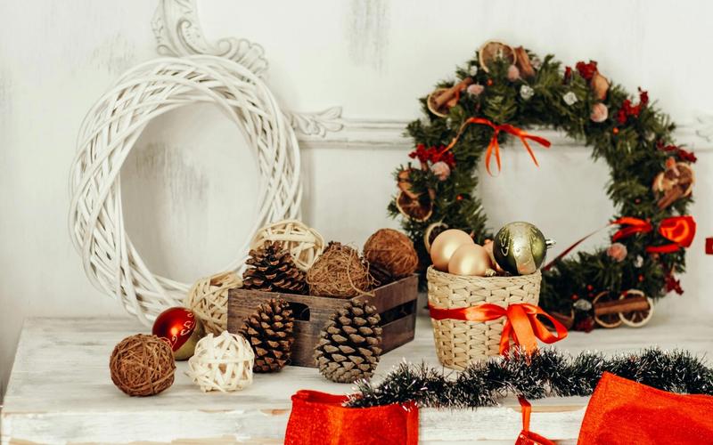 A white table decorated with pinecones, wicker balls, and Christmas ornaments in baskets evokes the charm of Gatlinburg Christmas, set next to a green holiday wreath with red ribbons and white wicker wreath, with red stockings hanging below.