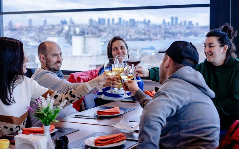 Five people sit around a table in a restaurant, smiling and clinking glasses for a toast, with a cityscape and water visible through large windows—perfect for large groups enjoying Gatlinburg dining.