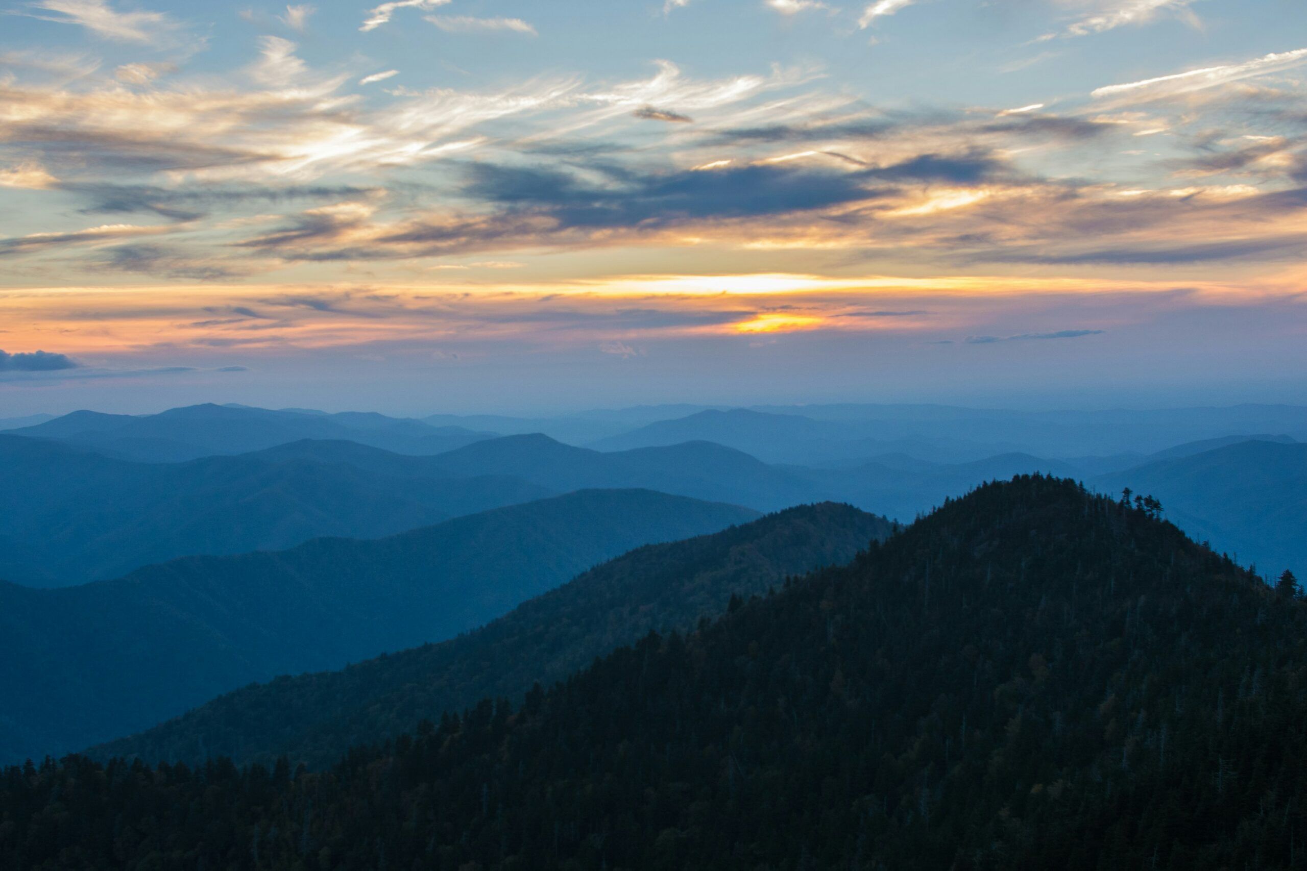 Sunset over a range of blue-tinted mountains near Gatlinburg, with layered ridges and a sky filled with scattered clouds and soft orange and yellow hues—a breathtaking scene among the top things to do in Gatlinburg.