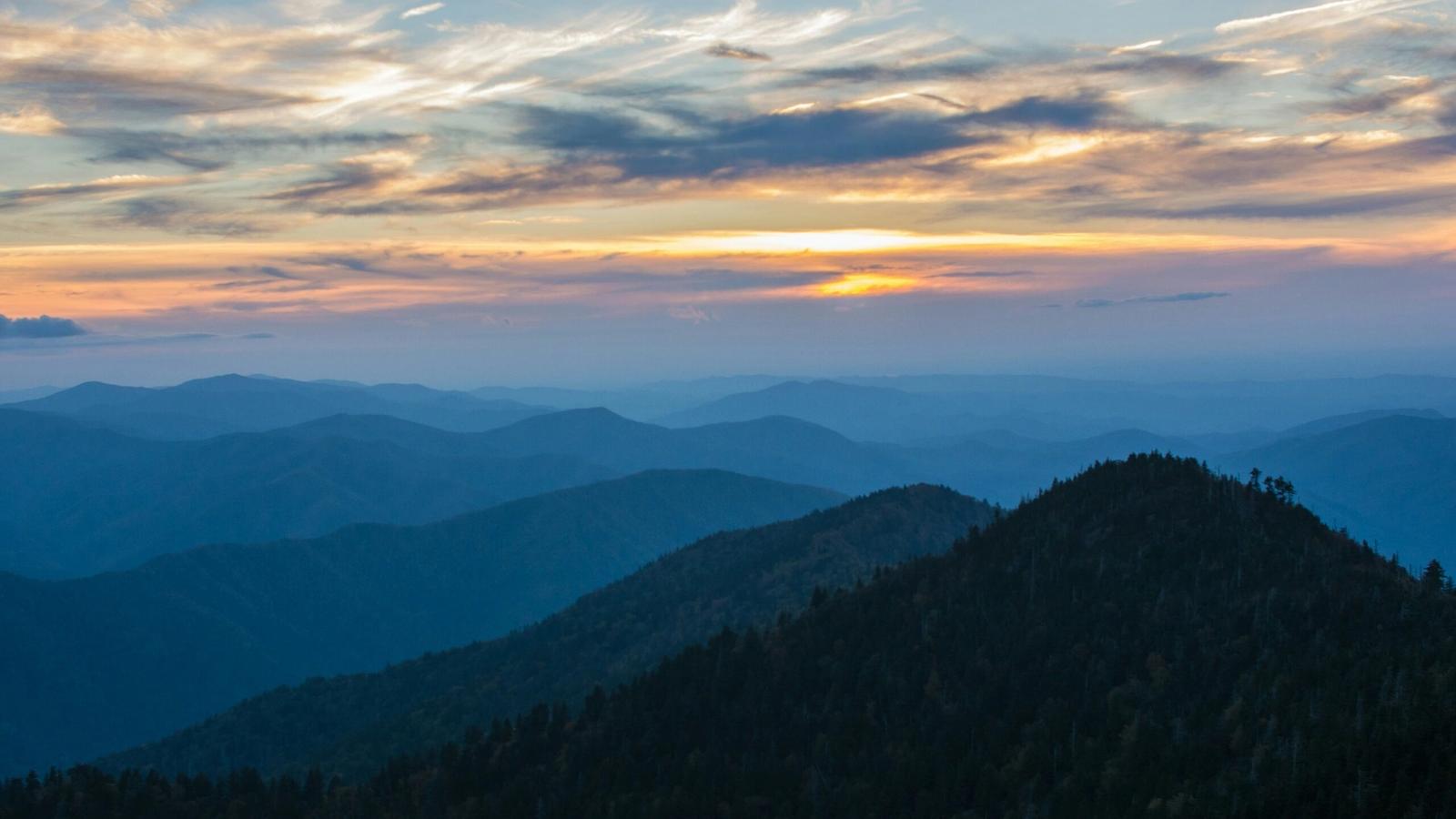 Sunset over a range of blue-tinted mountains near Gatlinburg, with layered ridges and a sky filled with scattered clouds and soft orange and yellow hues—a breathtaking scene among the top things to do in Gatlinburg.