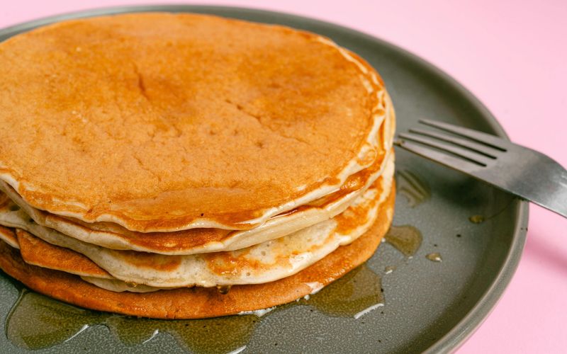 A stack of pancakes with syrup sits on a gray plate next to a fork, against a pink background, inspired by the menu at Crockett’s Breakfast Camp.
