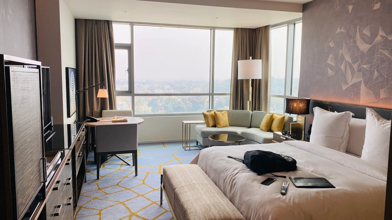 A modern luxury hotel room in Downtown Gatlinburg features a large bed, light-colored bench, sofa with yellow pillows, desk by the window, and cityscape views of the Strip. A TV, remote, and black bag are also visible in the room.