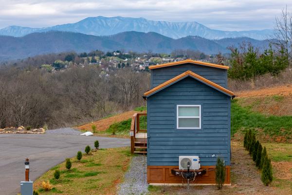 A small blue cabin sits on a grassy lot with the Smoky Mountain range and a town visible in the distance under a cloudy sky. Shrubs are planted along the path beside the house, creating a cozy atmosphere perfect for those who cherish close-knit communities.