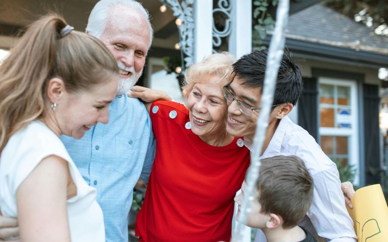 A group of five people, including two older adults, two younger adults, and a child, share a close, joyful hug outdoors in front of a house—smiling and enjoying each other’s company after playing Fun for Everyone family reunion games.