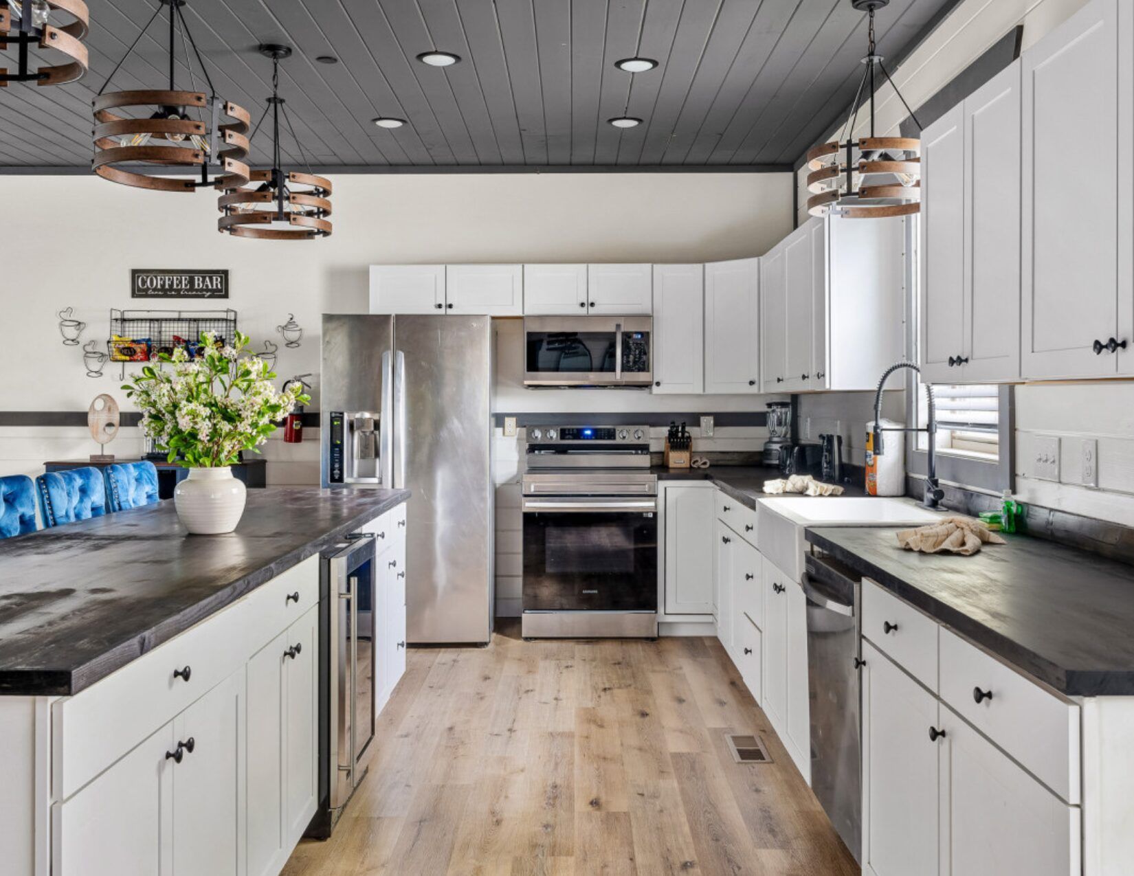 Modern kitchen with white cabinets, stainless steel appliances, black countertops, a farmhouse sink, wooden floor, and decorative light fixtures. A vase of flowers sits on the island, and a Coffee Bar sign is on the wall.