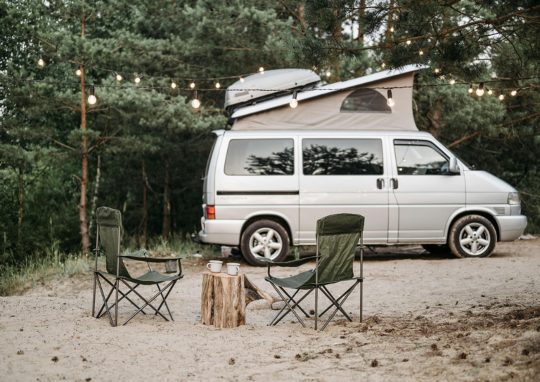 Two camping chairs and a small table with mugs sit on sandy ground in front of a campervan beneath hanging string lights, surrounded by pine trees—perfect for camping near Gatlinburg and enjoying the tranquility of the outdoors.