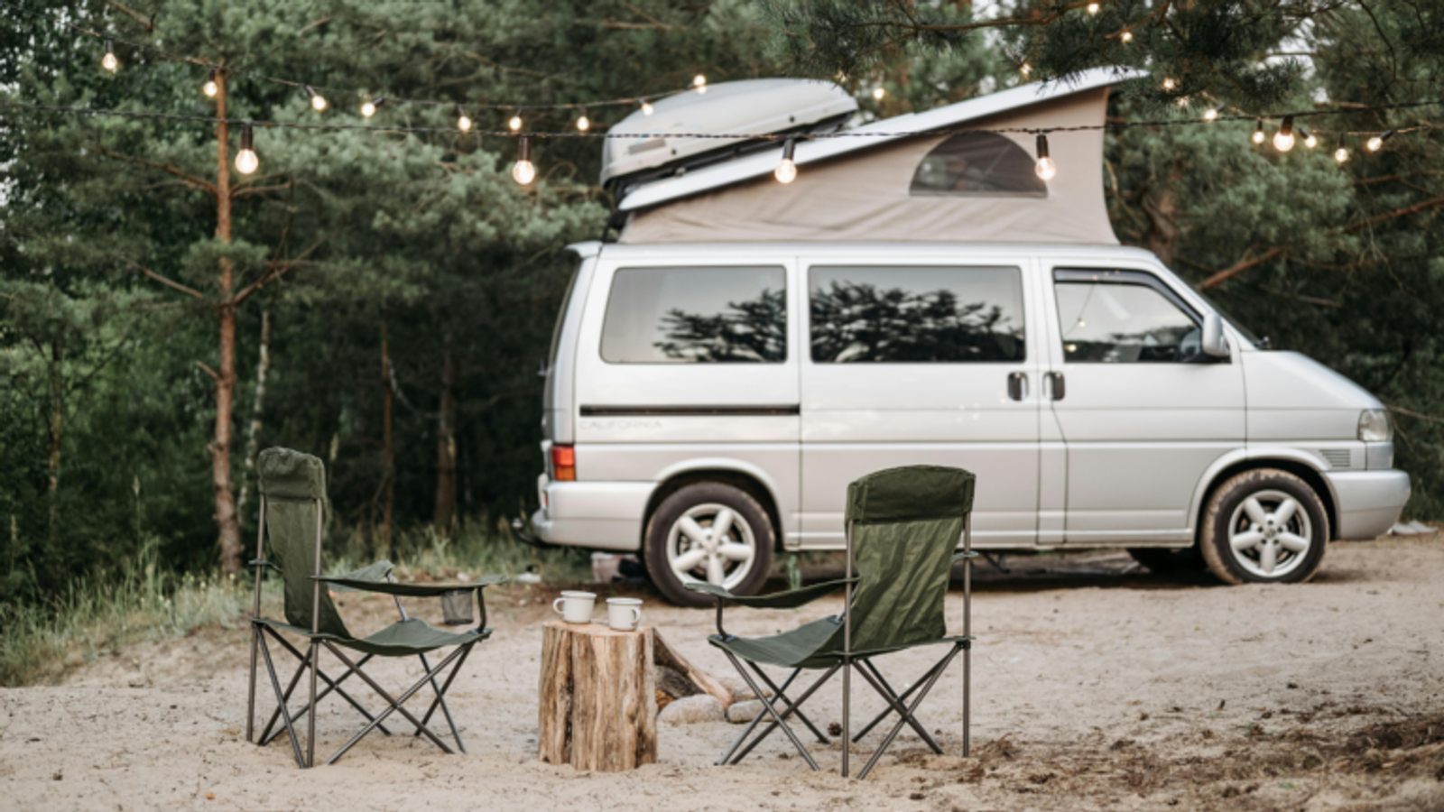 Two camping chairs and a small table with mugs sit on sandy ground in front of a campervan beneath hanging string lights, surrounded by pine trees—perfect for camping near Gatlinburg and enjoying the tranquility of the outdoors.