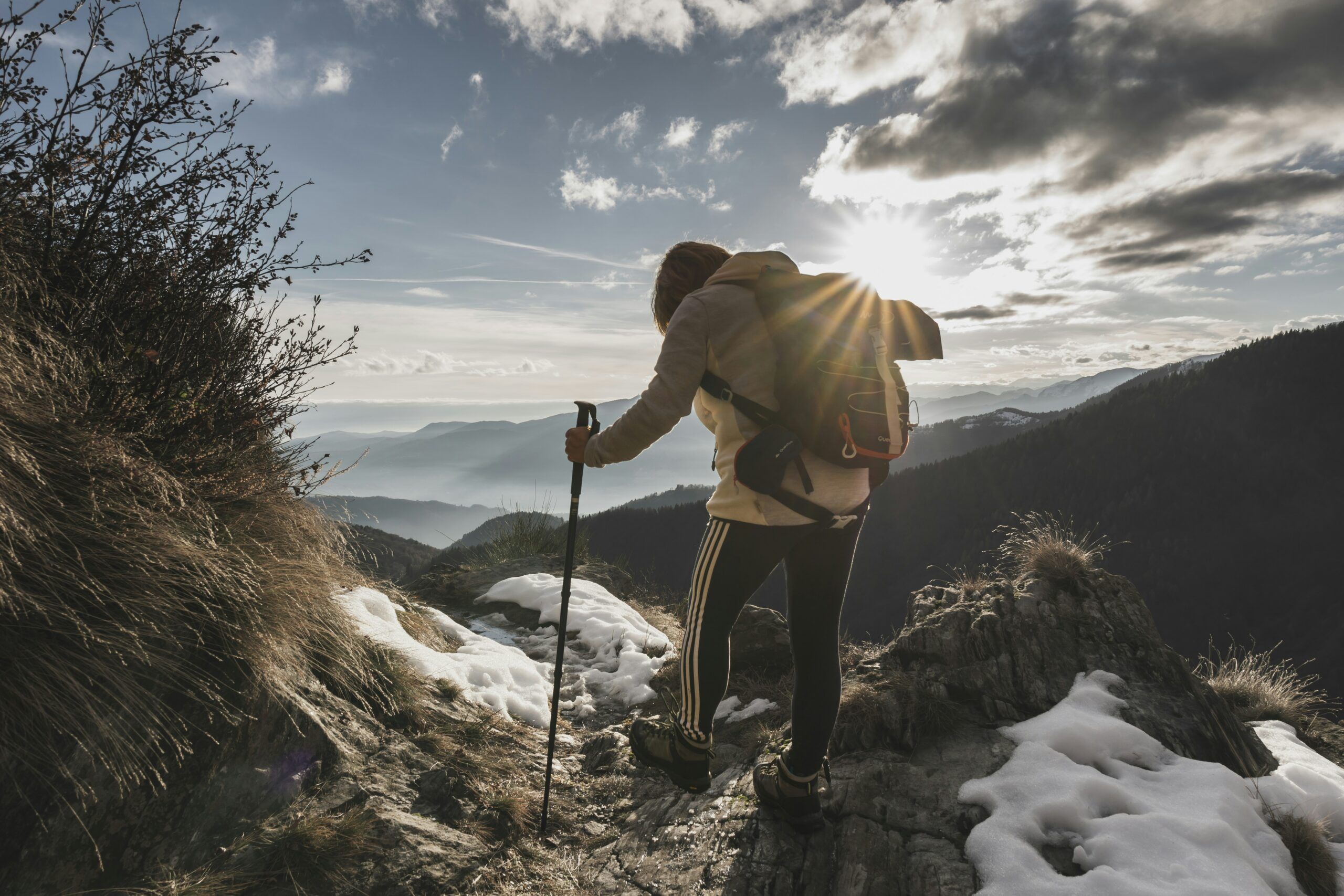 A hiker with a backpack and walking stick stands on a rocky, snow-dusted mountain slope at sunset, overlooking distant peaks and hiking trails in the Smoky Mountains under a partly cloudy sky.
