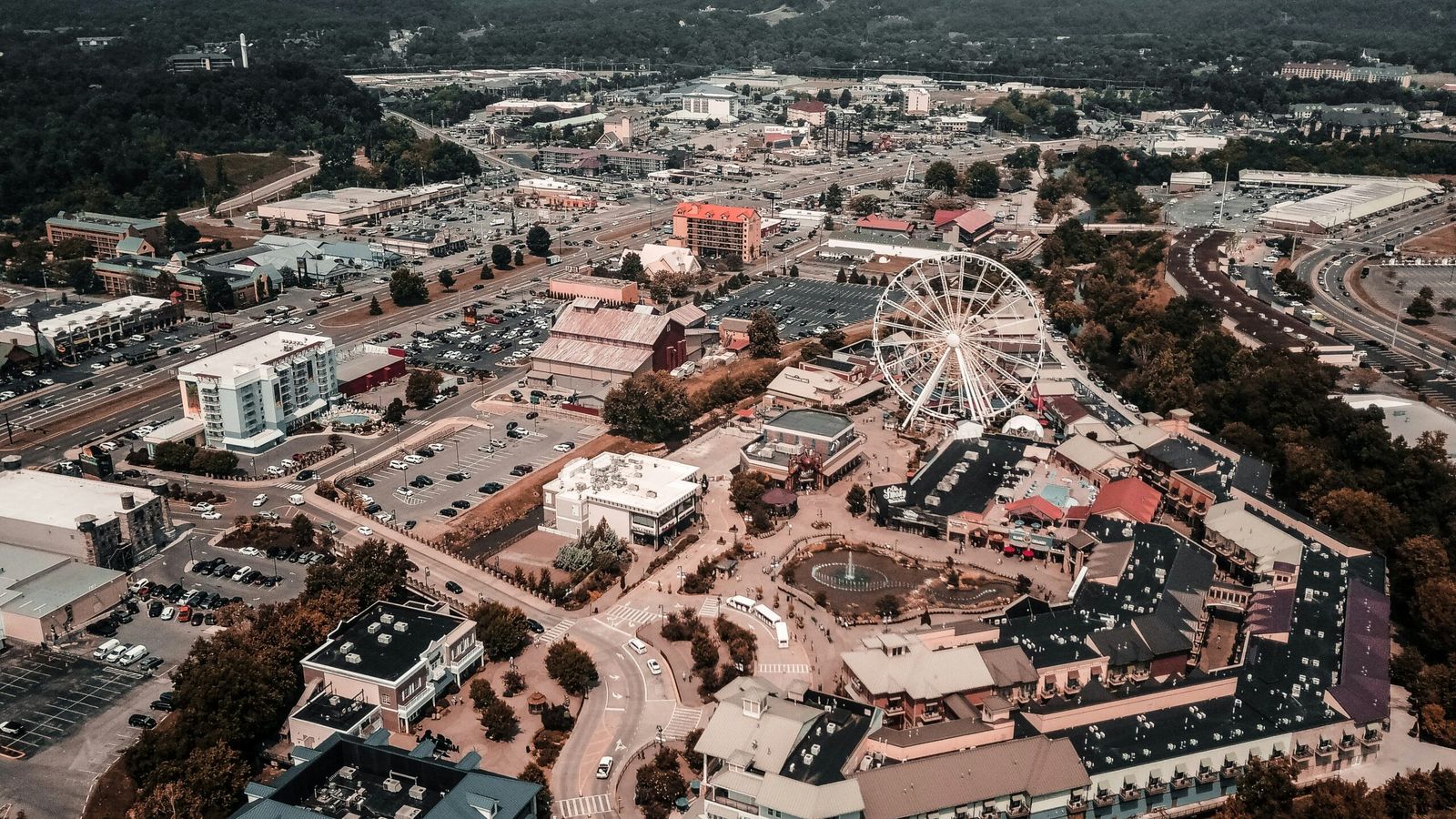 Aerial view of Pigeon Forge, showcasing its iconic Ferris wheel at the center, surrounded by city attractions, buildings, parking lots, roads, and distant green hills under a hazy sky.