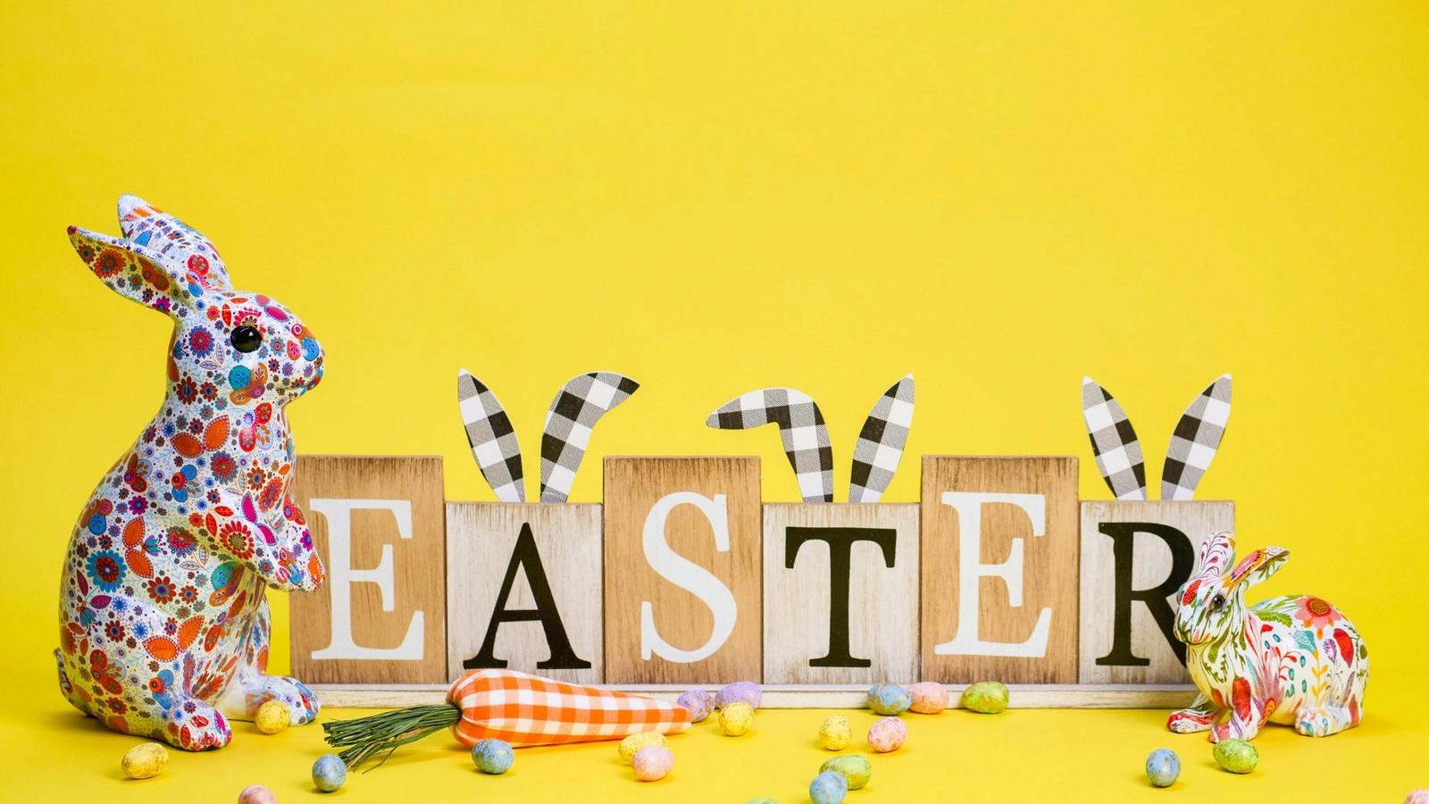 Colorful decorative rabbits and wooden blocks spelling EASTER with bunny ear decorations sit on a bright yellow background, surrounded by pastel candies and two fabric carrots—perfect inspiration for Smoky Mountains Easter activities.