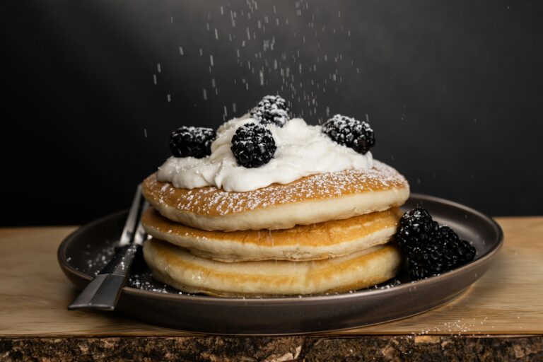A stack of pancakes topped with whipped cream and blackberries, being sprinkled with powdered sugar, sits on a dark plate—just like those you’ll find on the Log Cabin Pancake House menu in Gatlinburg TN. A fork rests beside it against a dark background.