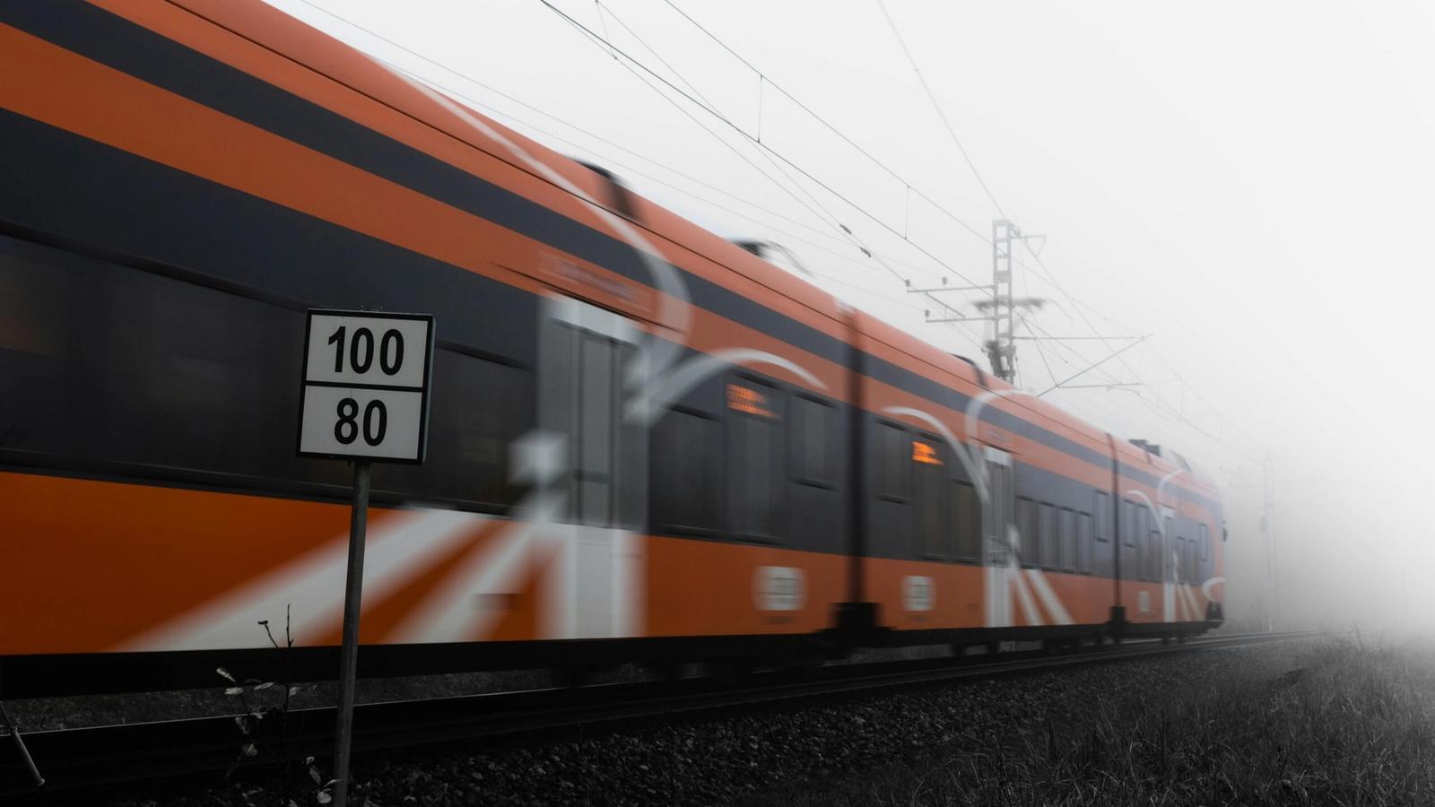 An orange train speeds past on railway tracks in a foggy setting, with a sign in the foreground displaying speed limits of 100 and 80—perfect inspiration for travel tips or things to do while exploring Gatlinburg in winter.