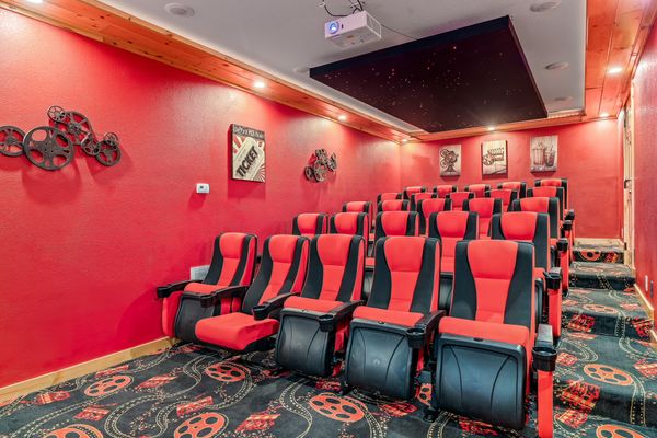 Home theater room with red walls, black and red reclining seats in rows, movie reel décor on the wall, framed movie-themed art, star-patterned ceiling panel, and patterned carpet with film reel designs.