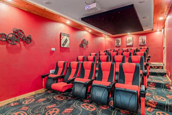Home theater room with red walls, black and red reclining seats in rows, movie reel décor on the wall, framed movie-themed art, star-patterned ceiling panel, and patterned carpet with film reel designs.