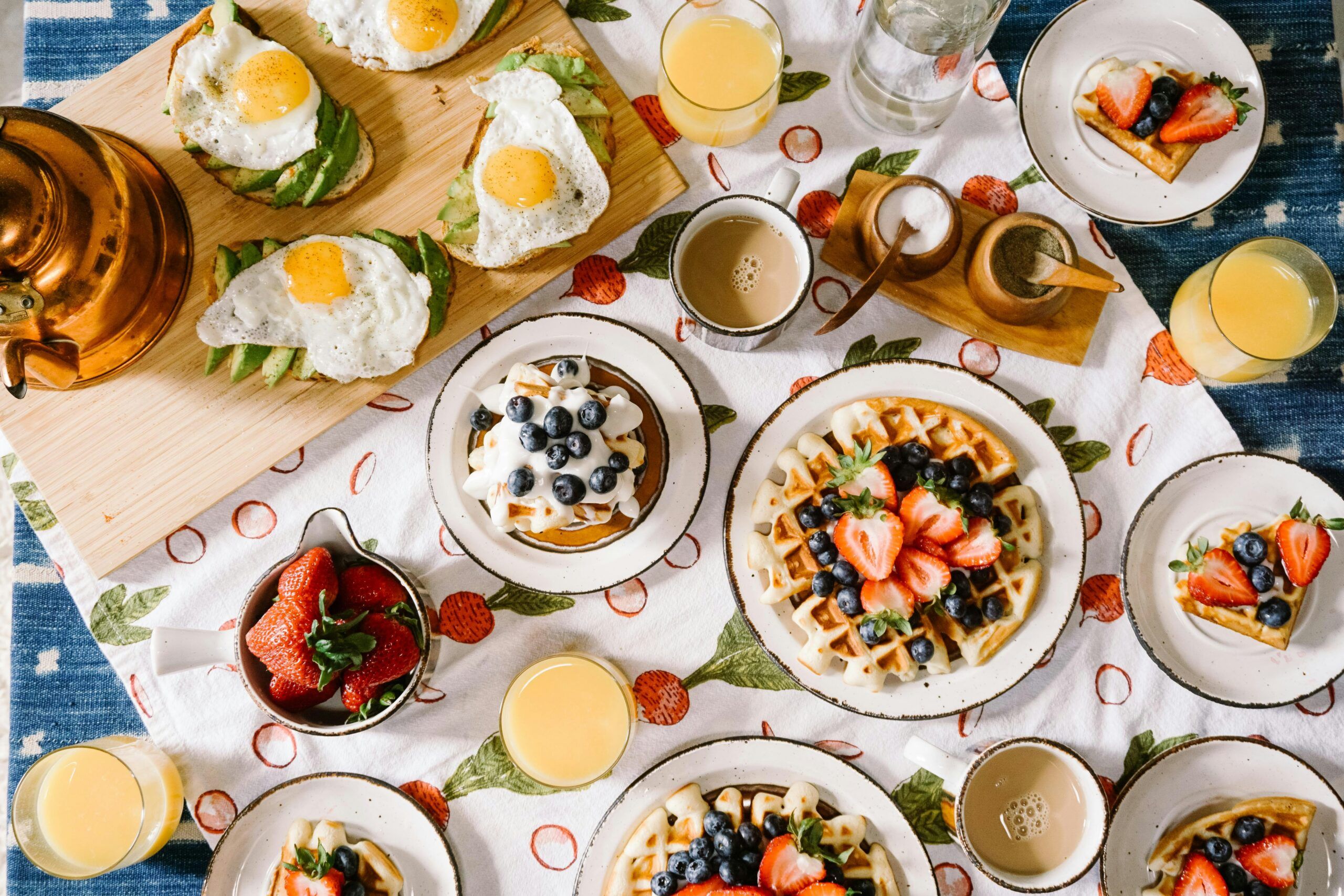 A breakfast table inspired by the best breakfast places in Gatlinburg, set with waffles topped with berries, avocado toast with fried eggs, orange juice, coffee, and a bowl of strawberries on a strawberry-patterned tablecloth.