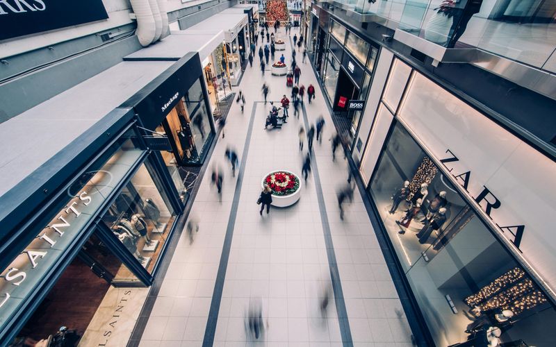 Aerial view of the busy Tanger Outlets Sevierville with blurred shoppers walking between store displays and clothing brand storefronts under bright lighting, perfect for vibrant shopping photos.