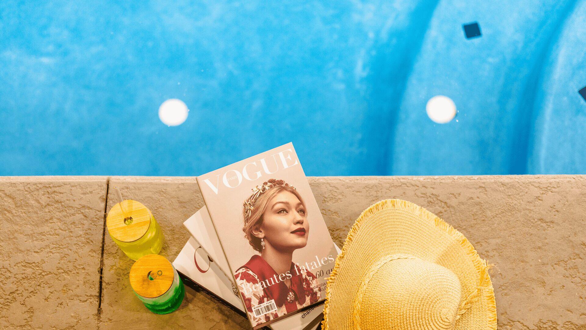 A Vogue magazine next to a pool, with a sun hat and two drinks.