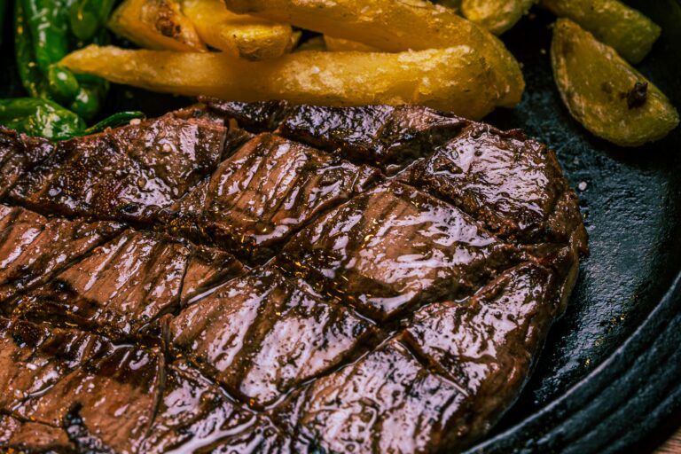 Close-up of a grilled steak with grill marks, served with golden French fries and green vegetables on a dark plate—just like you'd find on the Peddler Steakhouse menu. The steak appears juicy and cooked to perfection.