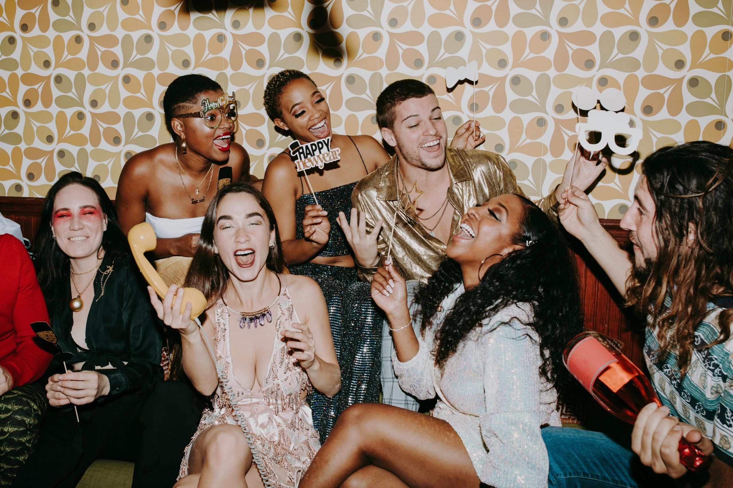 A group of friends dressed in festive outfits laugh and celebrate New Year's Eve together indoors in Gatlinburg, TN, holding party props and noisemakers against a retro-patterned wallpaper background.