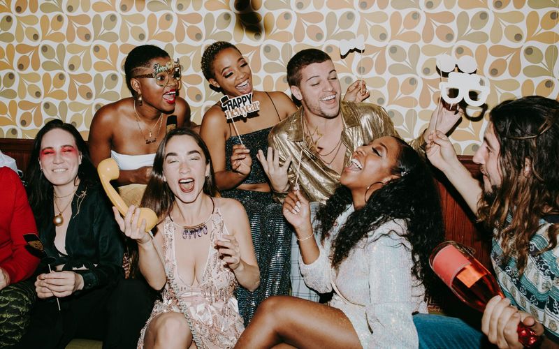 A group of friends dressed in festive outfits laugh and celebrate New Year's Eve together indoors in Gatlinburg, TN, holding party props and noisemakers against a retro-patterned wallpaper background.
