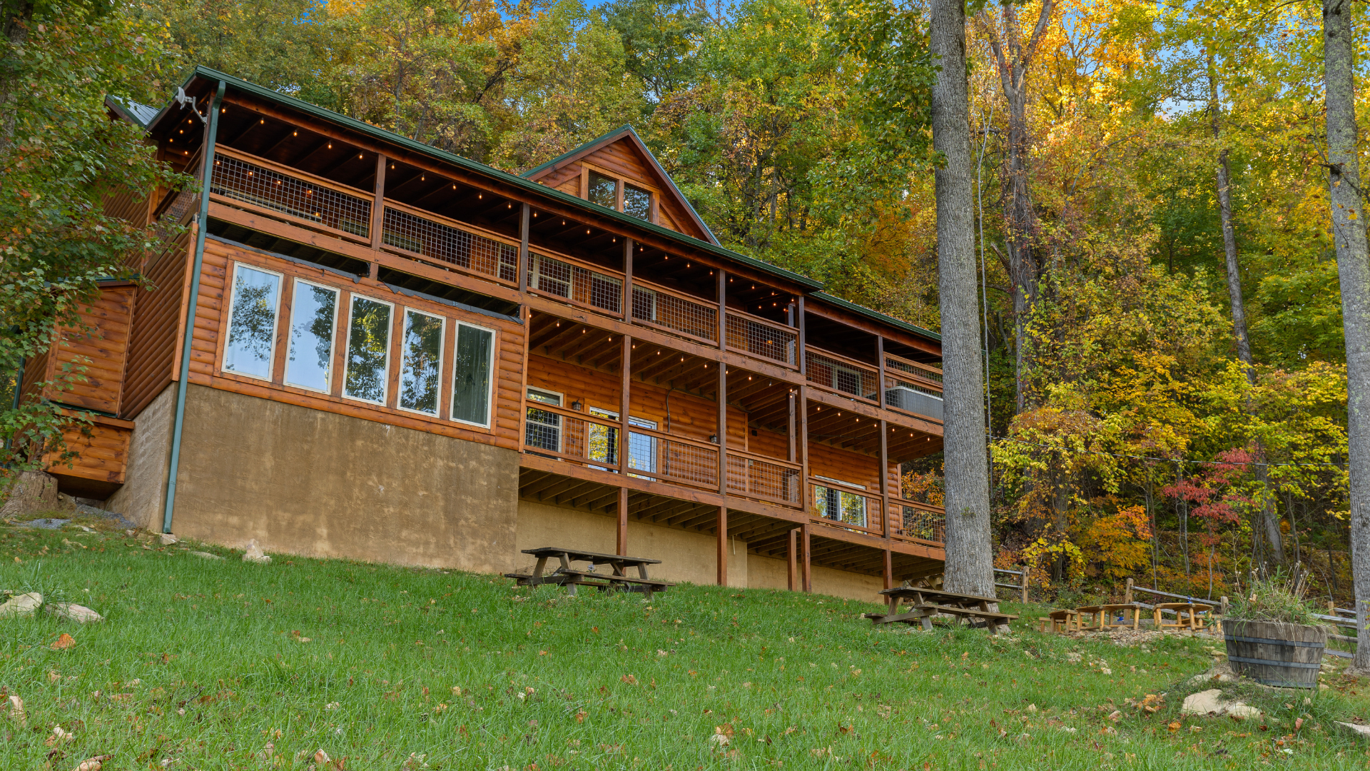 A large wooden cabin with a wraparound porch sits on a grassy slope, surrounded by tall trees with autumn foliage—perfect for wedding accommodations at Trillium Venue in the scenic Smoky Mountains. Picnic tables and a wooden barrel planter are in the yard.