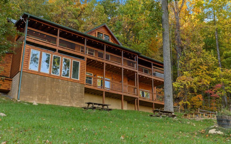 A large wooden cabin with a wraparound porch sits on a grassy slope, surrounded by tall trees with autumn foliage—perfect for wedding accommodations at Trillium Venue in the scenic Smoky Mountains. Picnic tables and a wooden barrel planter are in the yard.