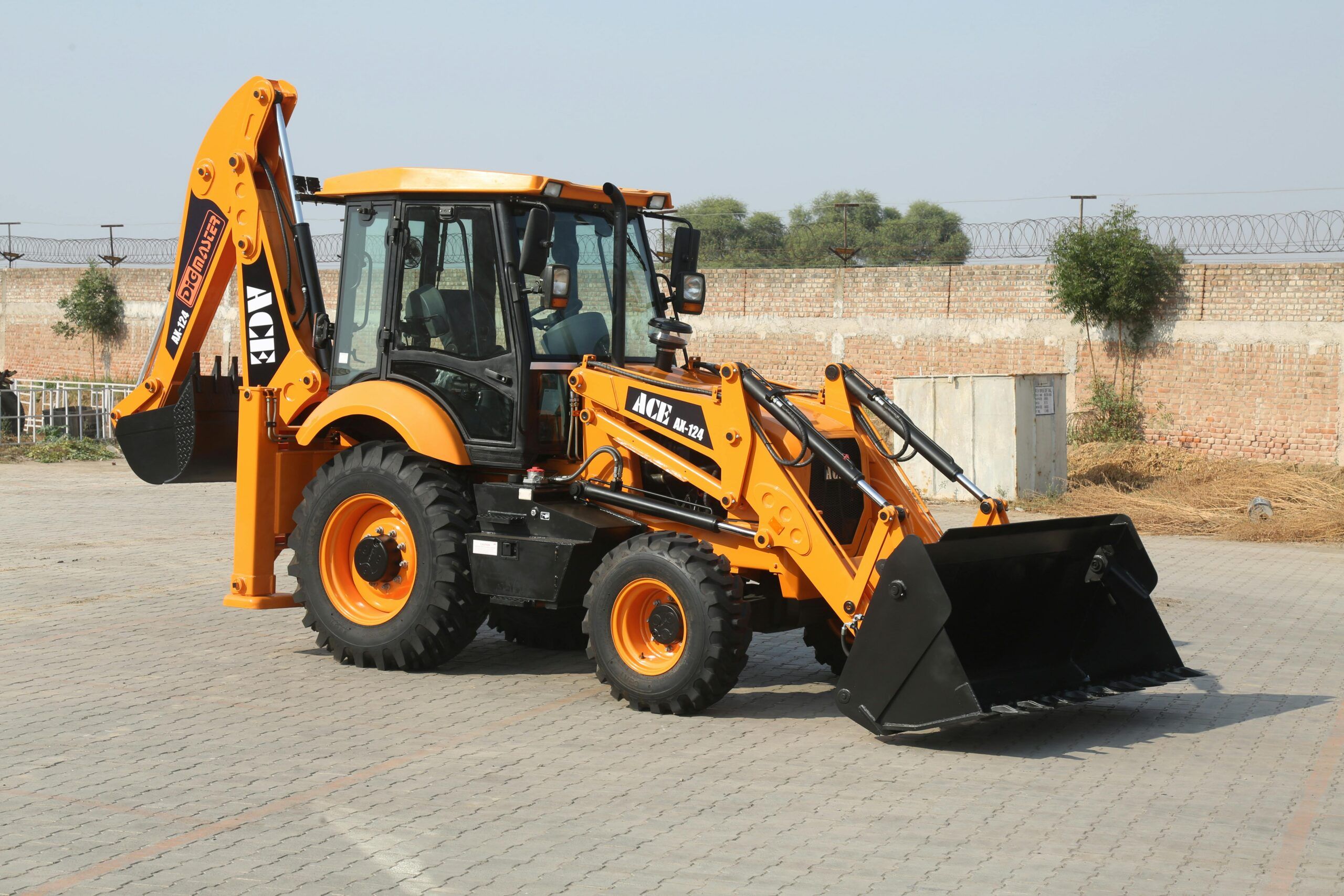 A yellow ACE backhoe loader is parked on a paved surface, featuring a front bucket and rear digging arm—just like the equipment you’ll see at Dig’n Zone Theme Park—with a brick wall and trees in the background.