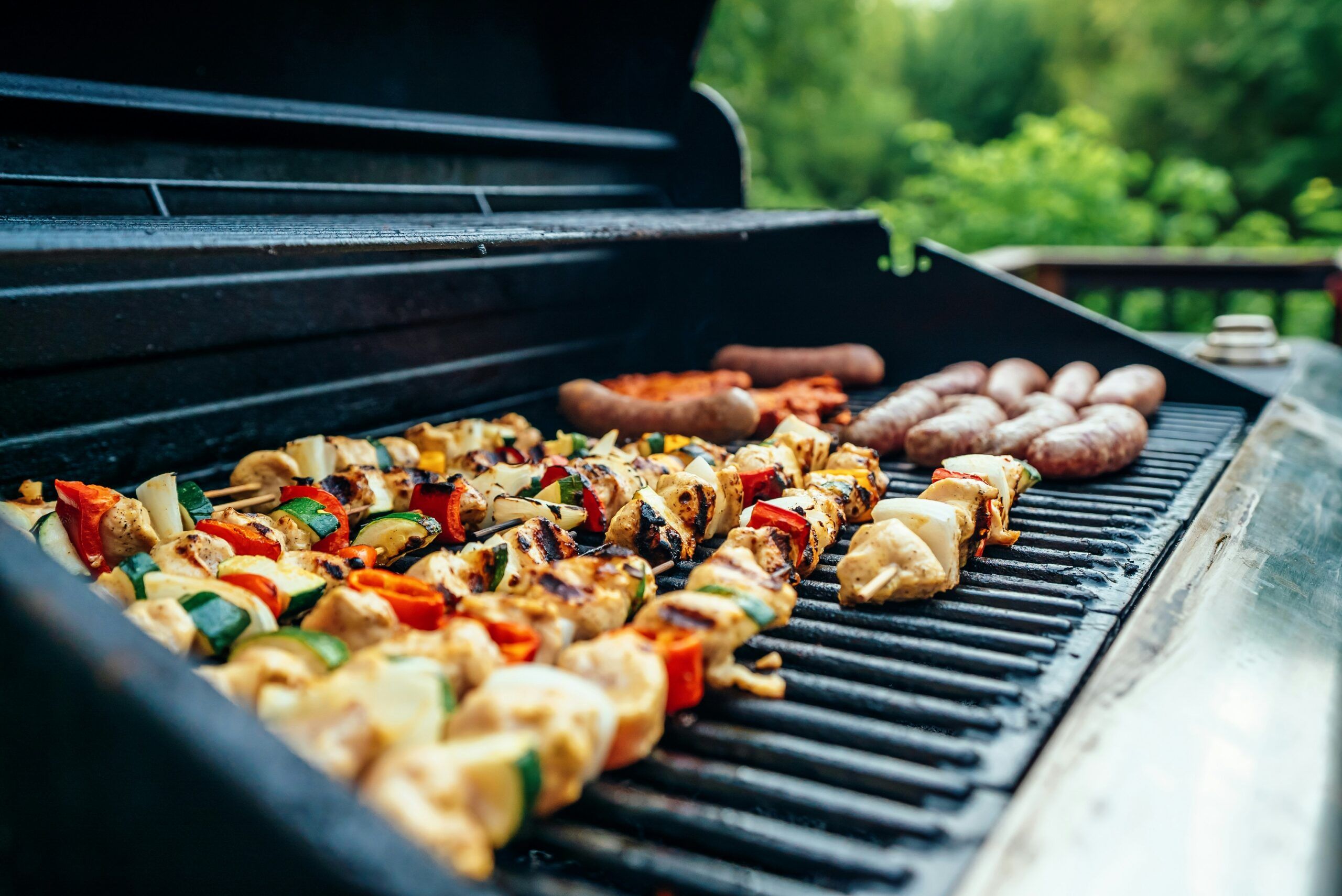 Colorful vegetable and chicken skewers, as well as sausages, sizzle on a Smokies BBQ, grilling outdoors with green trees visible in the blurred background.