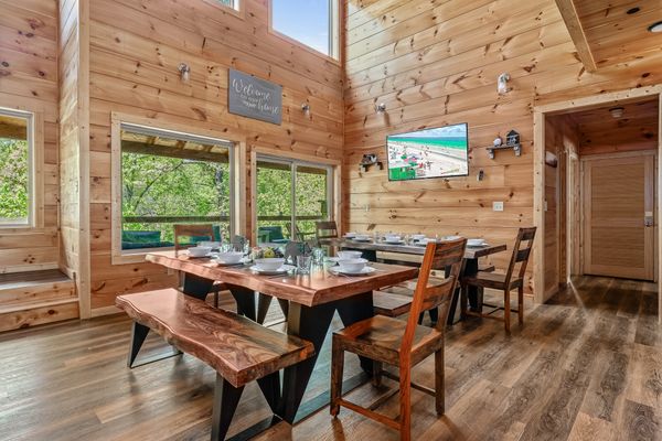 A cozy dining area with wooden walls and floors evokes the warm ambiance of Smoky Mountain cabins, featuring a large wooden table, bench, chairs, table settings, windows letting in natural light, and a TV mounted on the wall.