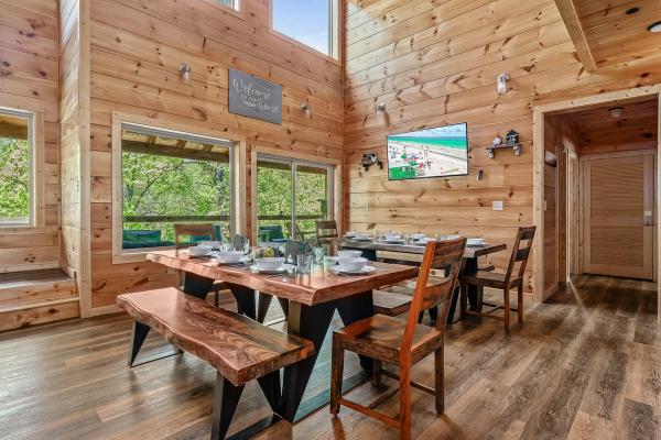 A cozy dining area with wooden walls and floors evokes the warm ambiance of Smoky Mountain cabins, featuring a large wooden table, bench, chairs, table settings, windows letting in natural light, and a TV mounted on the wall.
