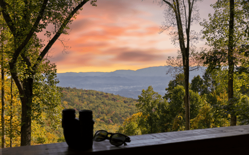 A scenic view of the Smoky Mountains and a colorful sunset sky, seen through tall trees. On a wooden ledge in the foreground are a pair of binoculars and sunglasses, perfect for relaxing after corporate retreats or enjoying local lodging.