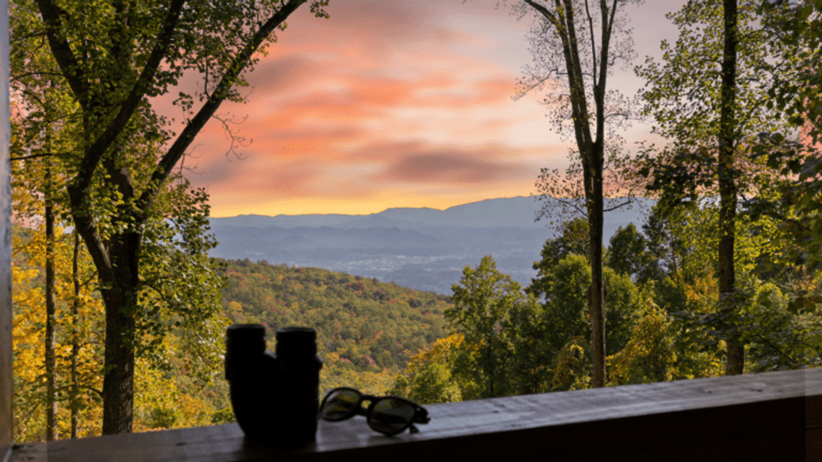 A scenic view of the Smoky Mountains and a colorful sunset sky, seen through tall trees. On a wooden ledge in the foreground are a pair of binoculars and sunglasses, perfect for relaxing after corporate retreats or enjoying local lodging.