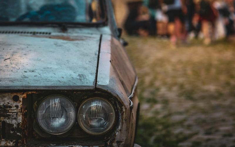 Close-up of the front left side of an old, rusty car with two round headlights, parked on a cobblestone road. The blurred background hints at people mingling, capturing the authentic vibe often found at Pigeon Forge car shows.