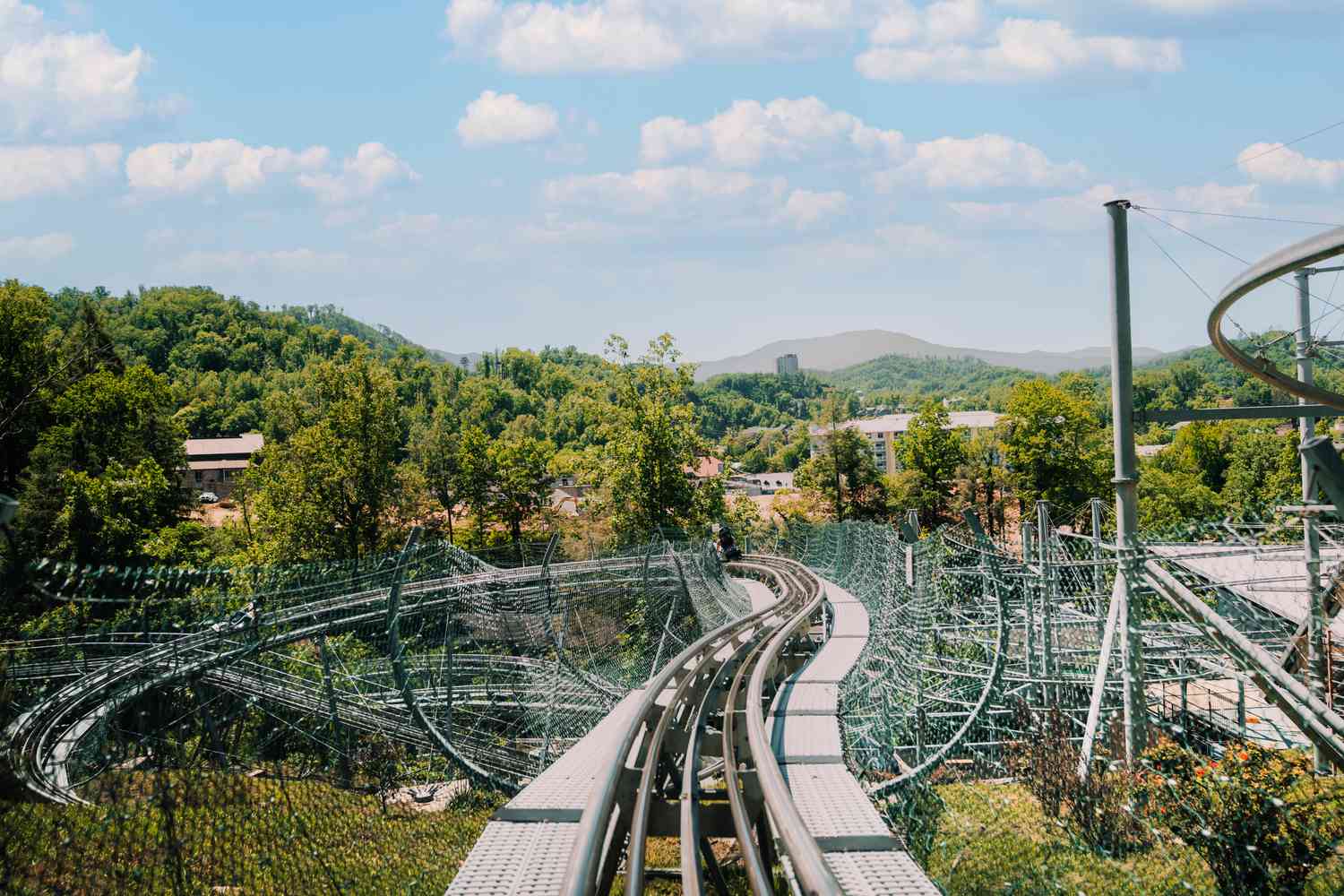 A view from the front seat of the Smoky Mountain Alpine Coaster track winding through green trees and hills, with blue sky and scattered clouds in the background.