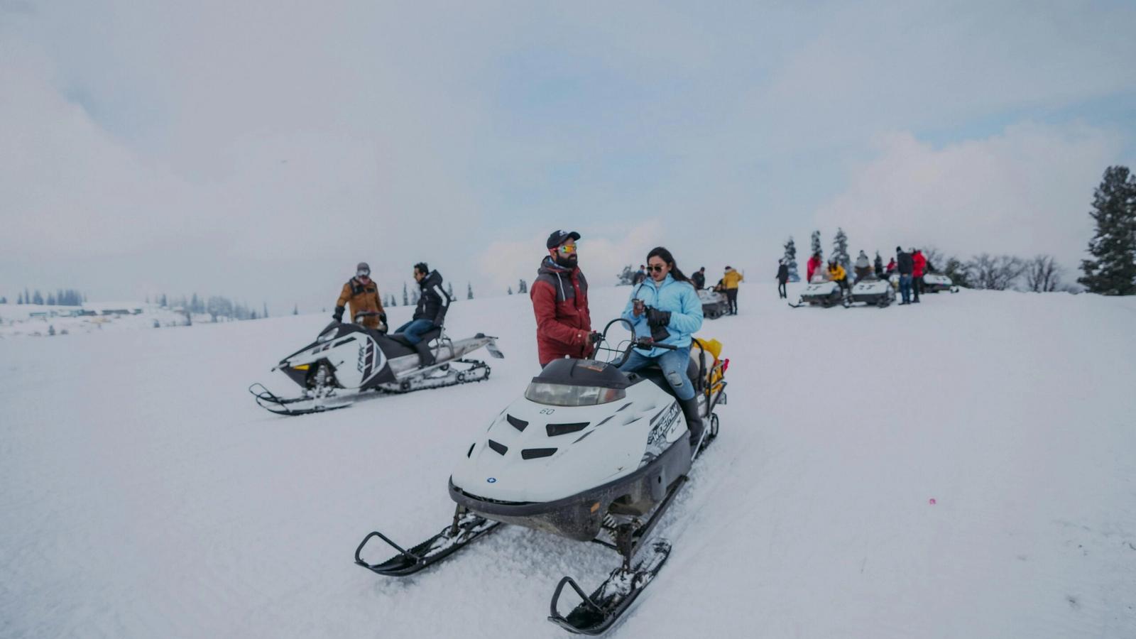 People enjoying winter activities on snowmobiles in a snowy landscape near Gatlinburg, TN. Dressed in winter clothing, some riders are moving while others are stationary among trees under a cloudy sky.