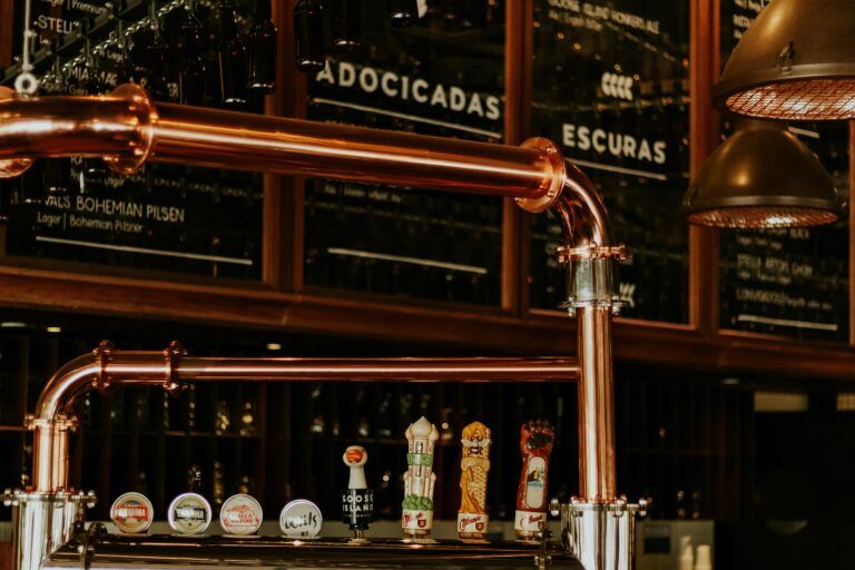 Beer taps with various labels are displayed on a polished copper bar in Gatlinburg. Glasses hang above, and a chalkboard menu with categories in Portuguese is visible in the background under warm lighting, evoking local distilleries.