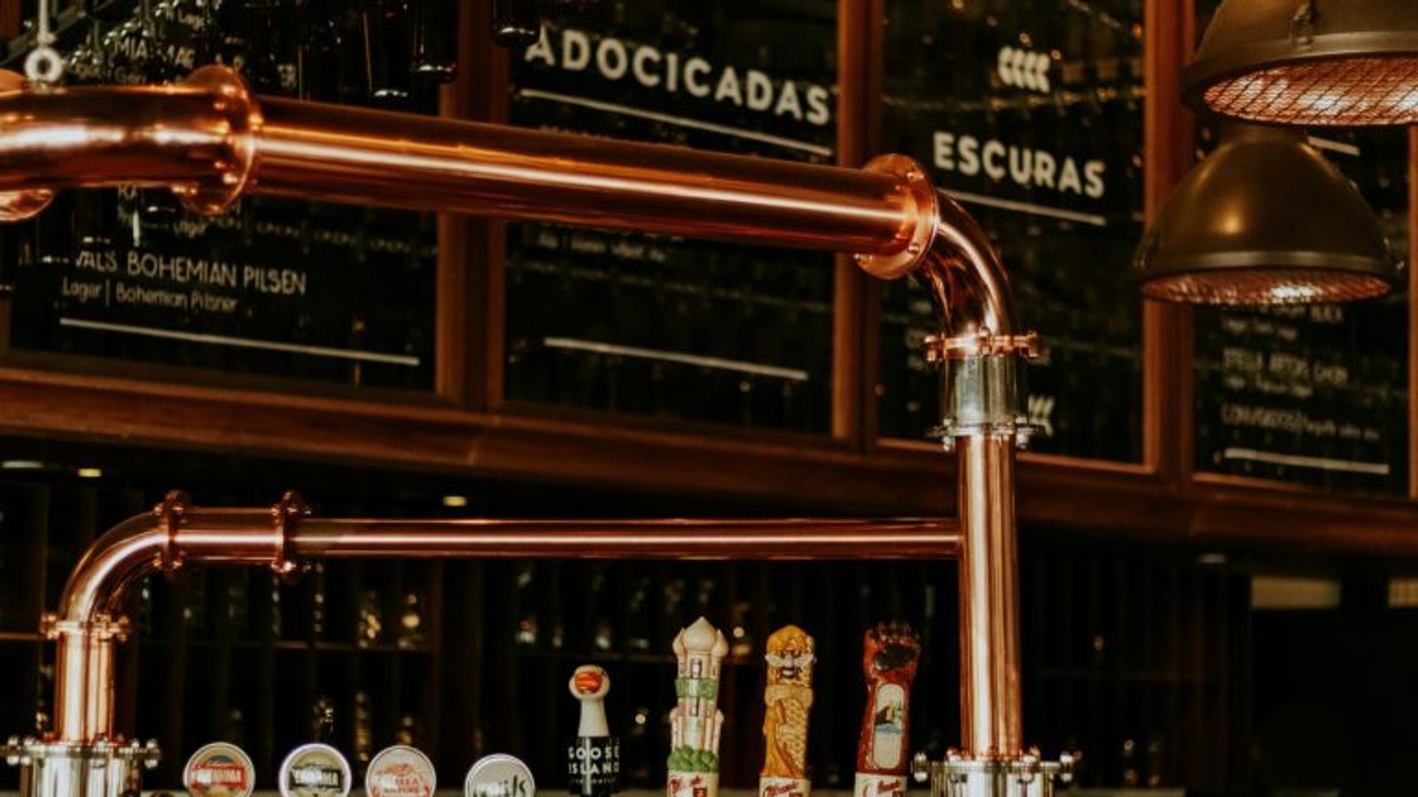 Beer taps with various labels are displayed on a polished copper bar in Gatlinburg. Glasses hang above, and a chalkboard menu with categories in Portuguese is visible in the background under warm lighting, evoking local distilleries.