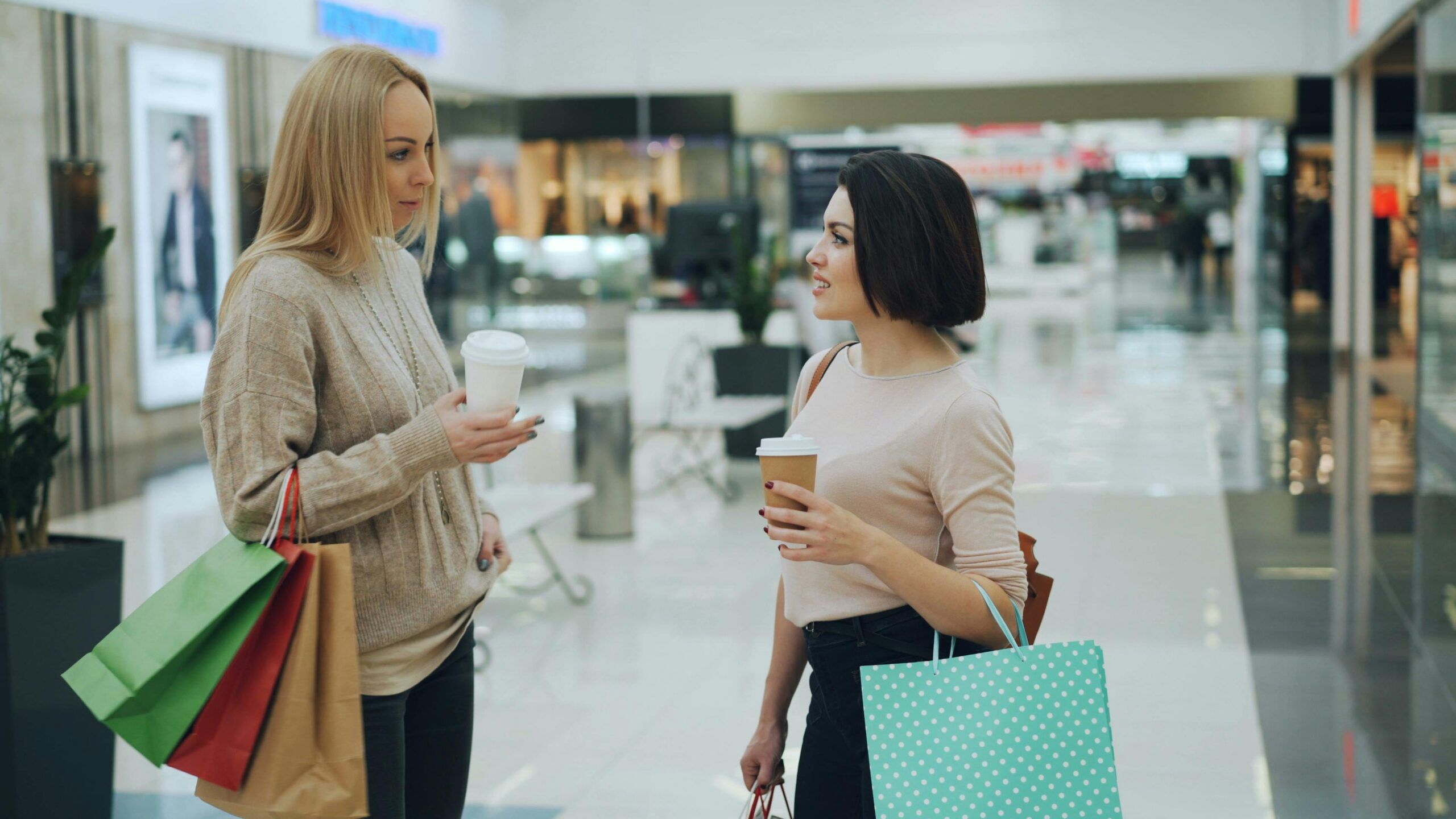 Two women holding shopping bags and coffee cups stand and talk in a brightly lit mall, surrounded by stylish shops in Gatlinburg. One woman has blonde hair and a beige sweater; the other has dark hair and a pale top. Storefronts and displays are visible behind them.