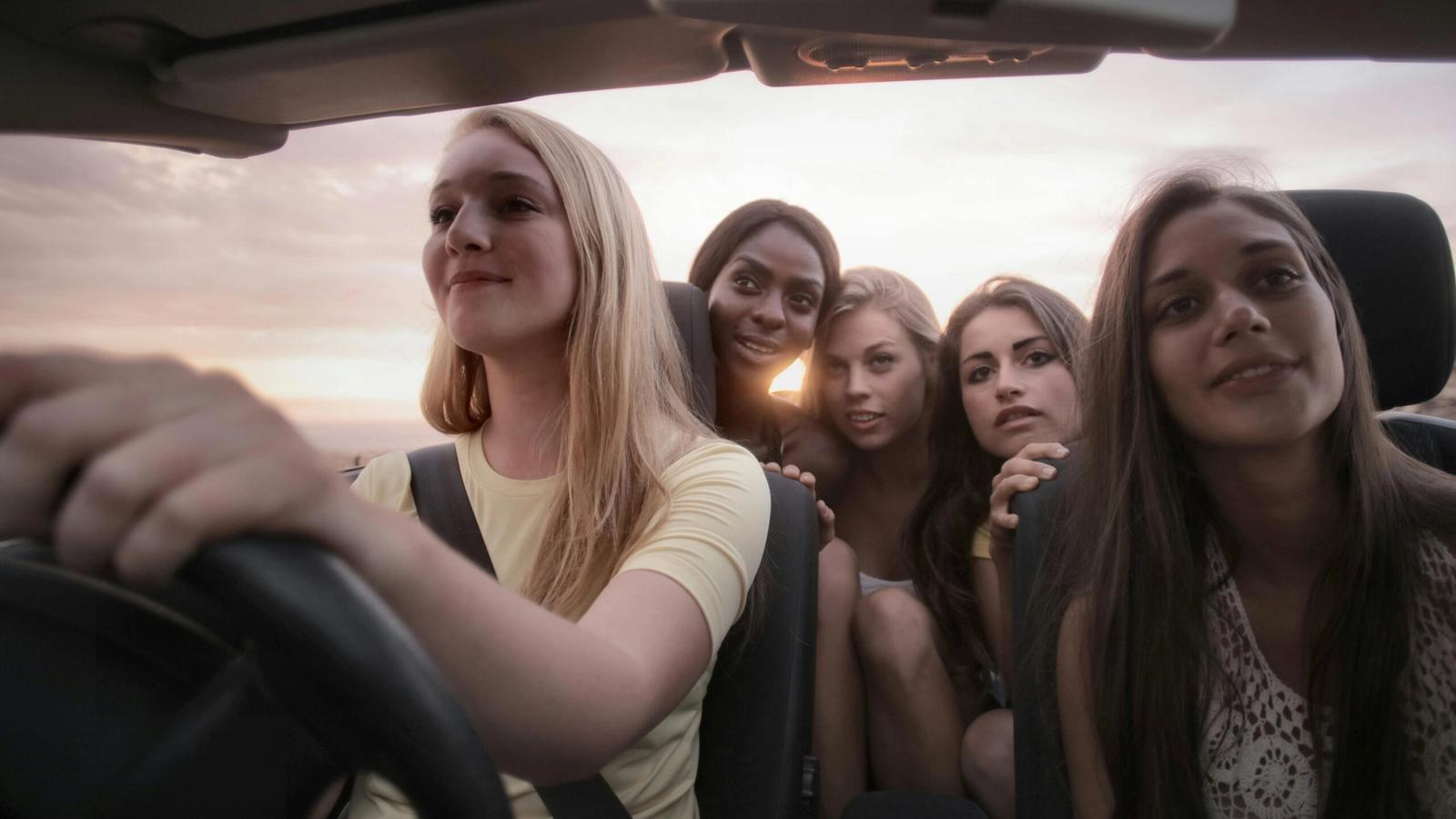 Five young women sit together in a car at sunset, with one driving and the others smiling and posing for a photo, capturing the cheerful spirit of a Girls’ Trip as they head toward one of the Best Places on their journey.