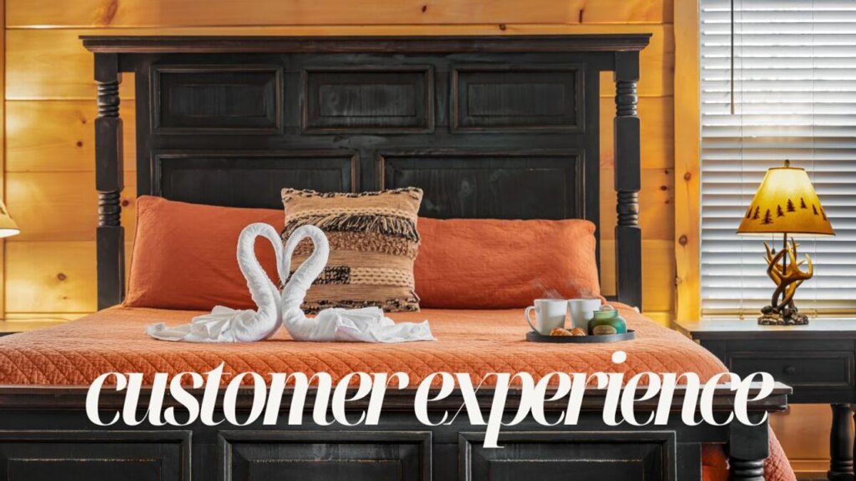 A cozy bedroom with an orange bedspread, a decorative pillow, and two towel swans forming a heart. Breakfast and coffee sit on a tray. The words Why We Stand Out are overlaid at the bottom.
