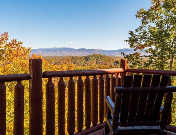 A wooden chair sits on a porch with a log railing, overlooking a scenic view of tree-covered hills and distant mountains under a clear blue sky.